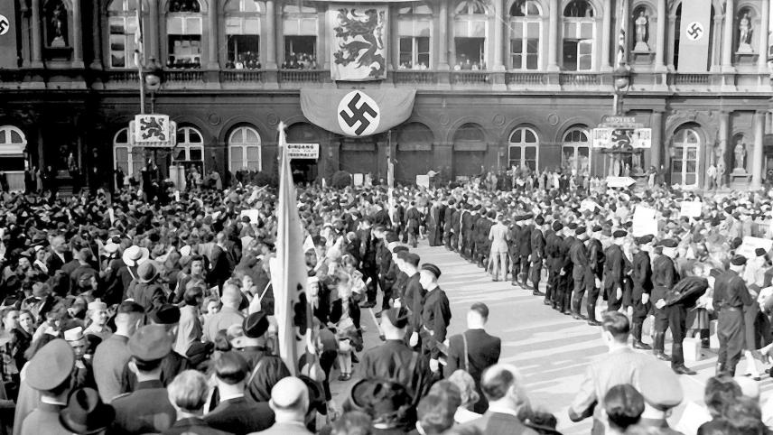 Le 2 septembre 1941, devant la gare du Nord, sur la place Rogier à Bruxelles, encadrées par les miliciens de DeVlag, des familles belges attendent leurs enfants qui ont passé leurs vacances en Allemagne.