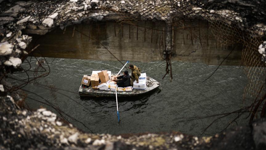 Un homme utilise son bateau plein de marchandises pour les habitants, avec un militaire ukrainien. Ils traversent la rivière Siverskyi-Donets, sous un pont détruit en Ukraine.